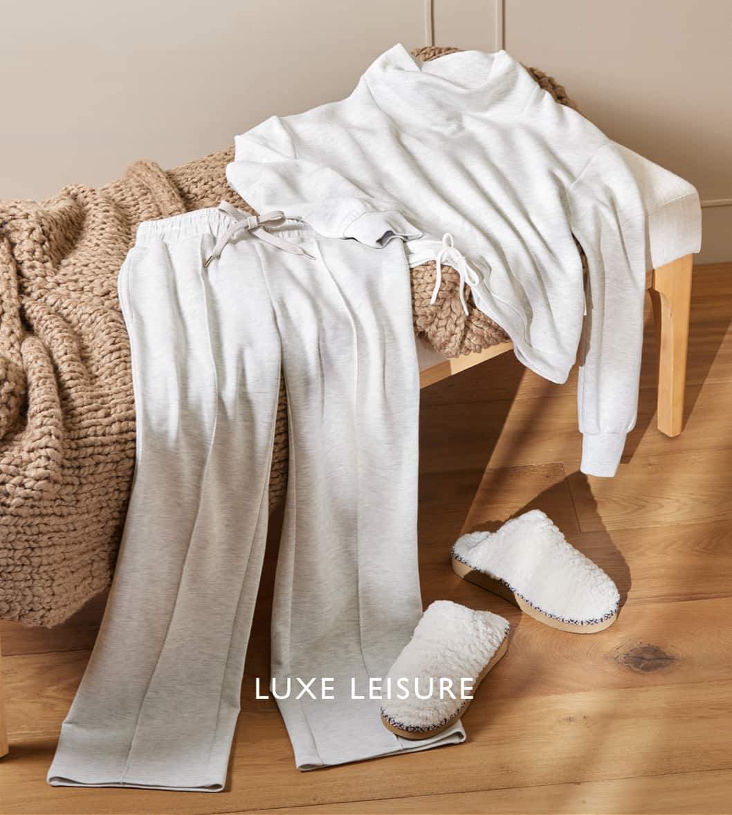 Image of an oatmeal heather colored sweatshirt and sweatpants laid on a bench with cream shearling slipper clogs on the floor with the caption "Luxe Leisure"